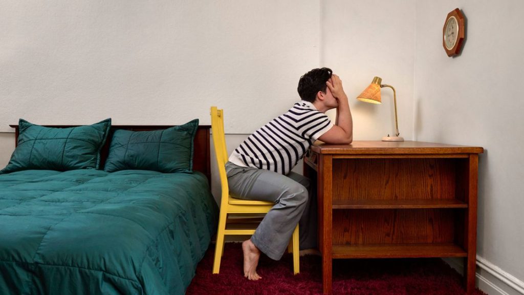 Man sitting at a bedroom desk holding his head, representing the discomfort of rectal itching and the search for relief
