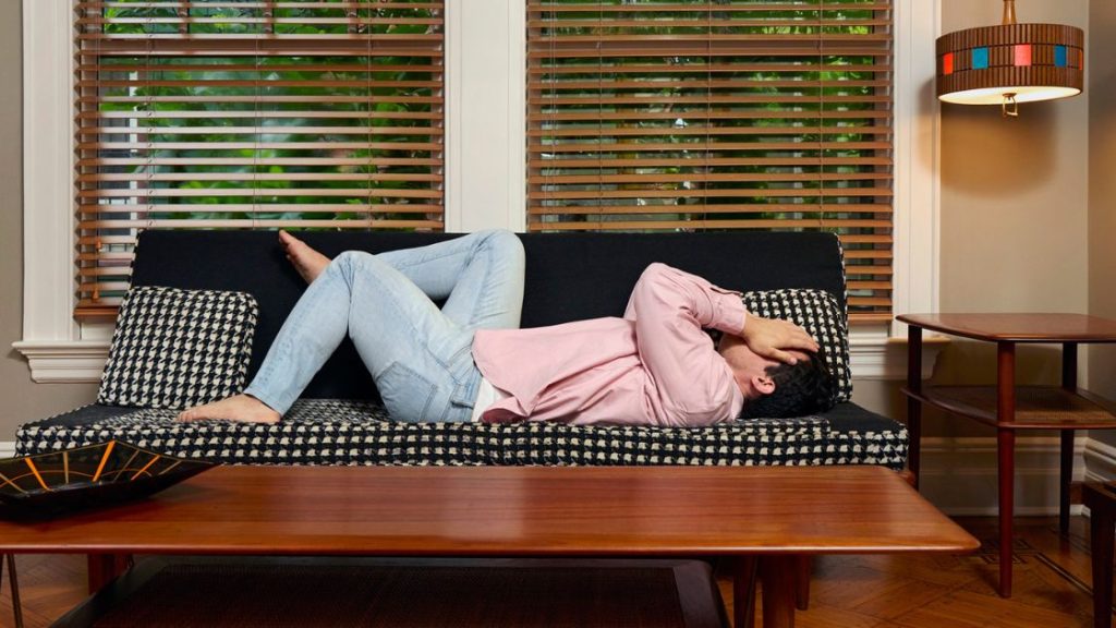Man curled up uncomfortably on a couch, holding his head, representing the distraction and discomfort of itchy anus symptoms